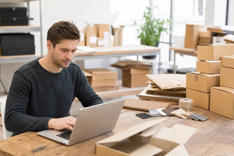 Onlinehändler prüft am Laptop Verpackungsmengen neben Versandkartons und Materialproben