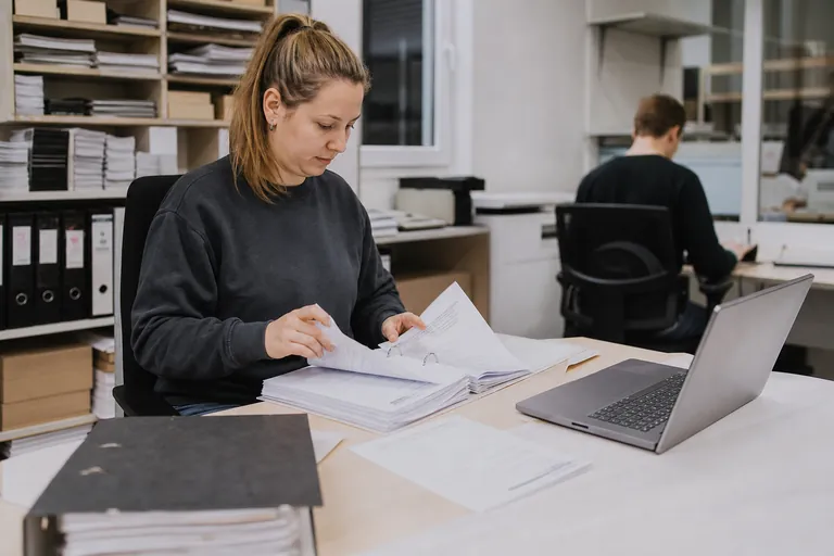 Sortierte Unterlagen zu Verpackungsmengen liegen neben Laptop und Ordner auf einem Schreibtisch